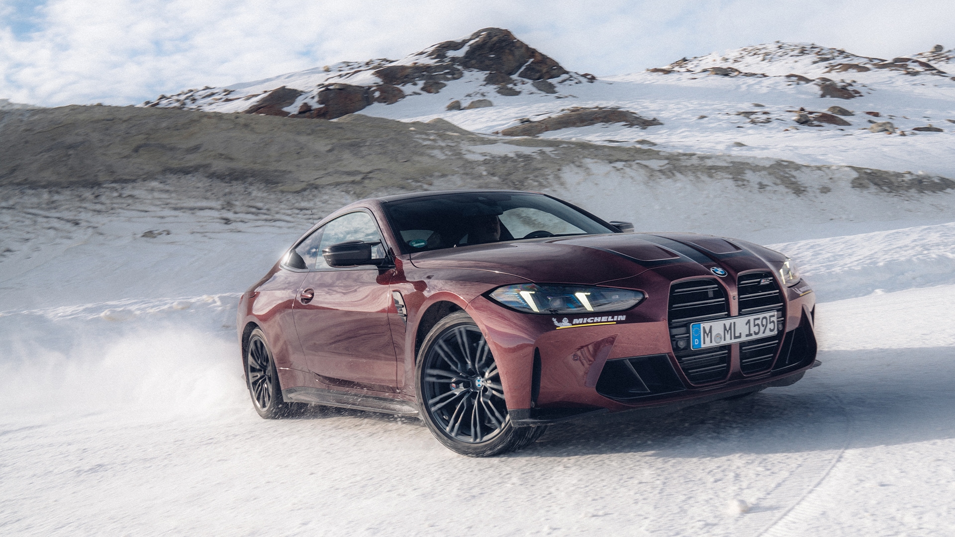 Ein burgunderroter BMW Sportwagen fährt durch eine verschneite Landschaft mit schneebedeckten Bergen im Hintergrund. Das Fahrzeug hat markante schwarze Räder und den charakteristischen BMW-Kühlergrill, mit einem sichtbaren Kennzeichen an der Front.