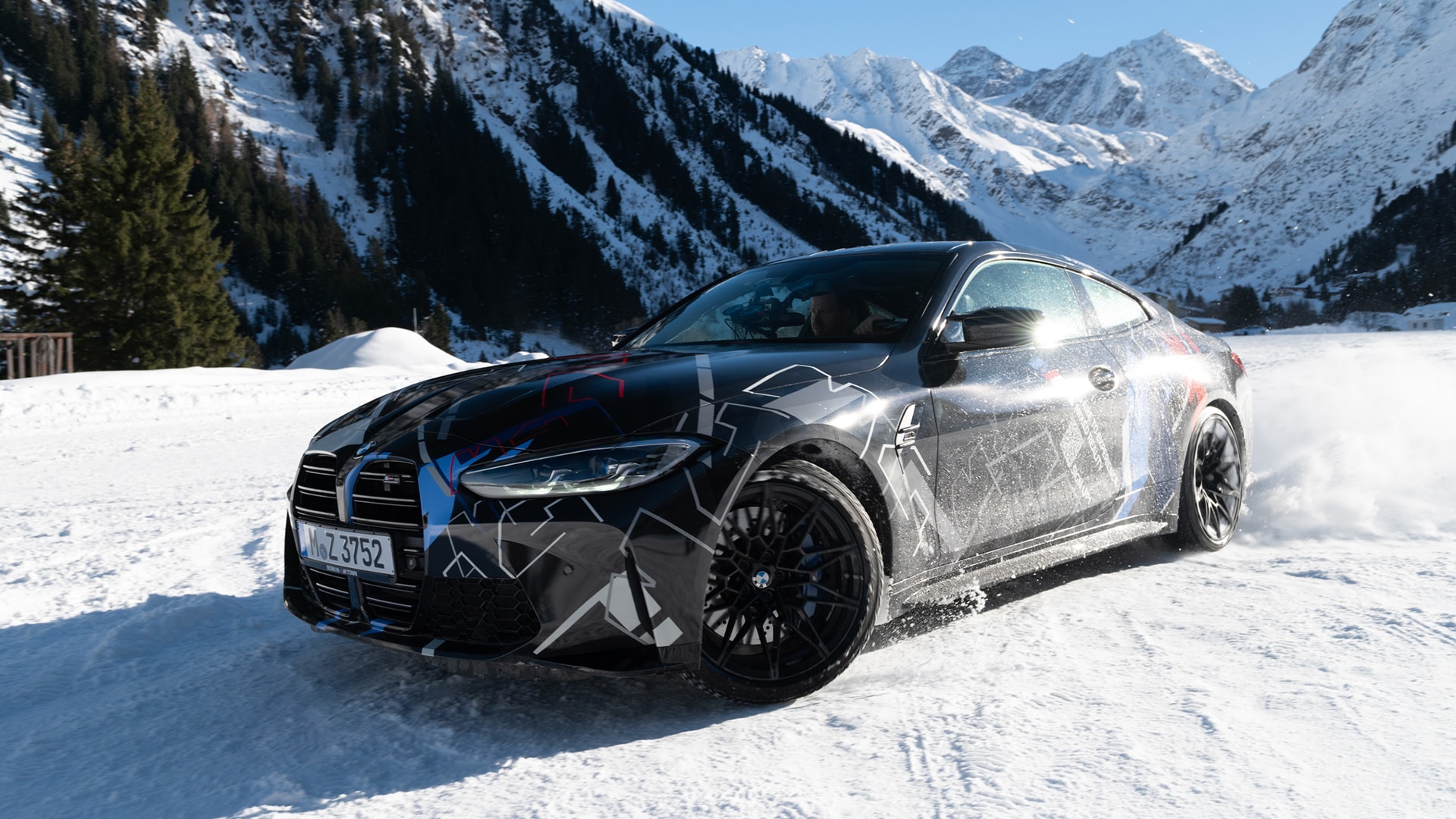 Ein schwarzer BMW Sportwagen mit Tarnmuster-Folierung fährt durch schneebedecktes Gelände. Das Fahrzeug steht vor einer beeindruckenden Kulisse aus schneebedeckten Bergen in einer alpinen Umgebung mit immergrünen Bäumen auf der linken Seite.