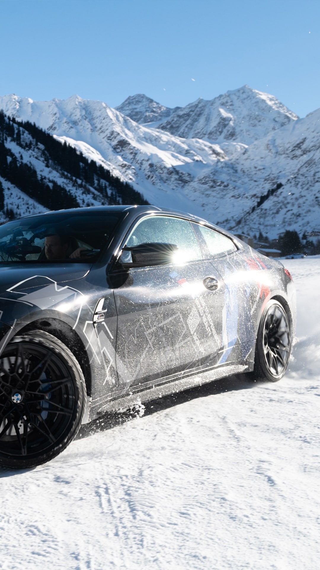 Ein schwarzer Sportwagen fährt auf einer schneebedeckten Straße, wobei Schnee von den Rädern spritzt. Im Hintergrund erheben sich majestätische schneebedeckte Berge gegen einen klaren blauen Himmel und bilden eine dramatische Alpenlandschaft.