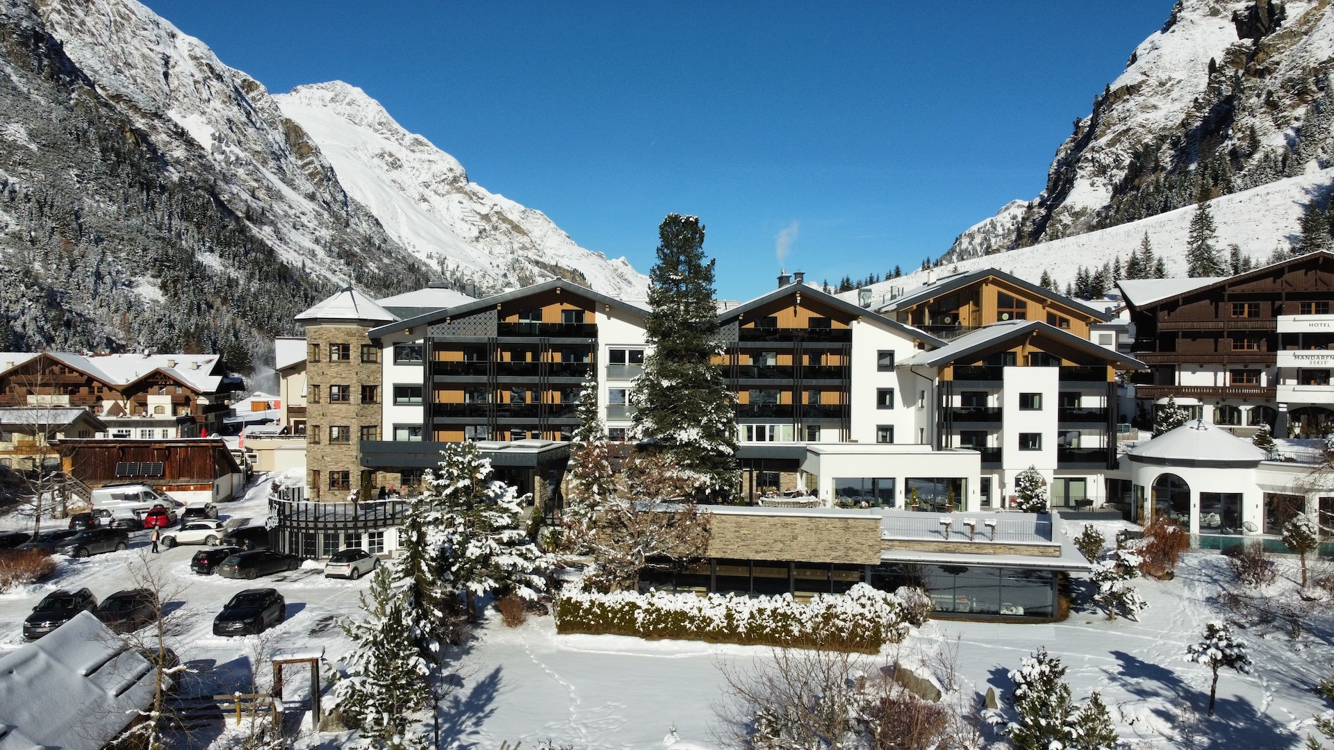 Ein winterliches Alpenresort in einem schneebedeckten Tal mit mehreren Chalets und Hotelbauwerken. Schneebedeckte Berge erheben sich dramatisch zu beiden Seiten unter einem klaren blauen Himmel. Im Vordergrund ist ein Parkplatz mit mehreren Autos zu sehen, alle mit Schnee bedeckt. Immergrüne Bäume säumen die Landschaft um die Gebäude.
