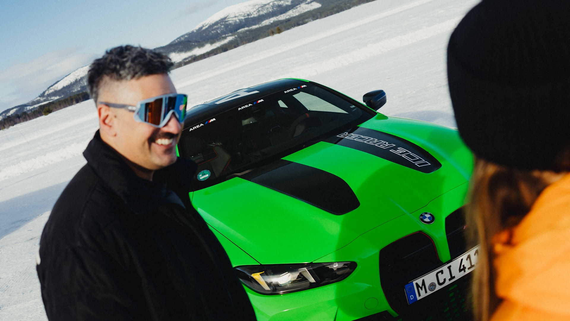 Lächelnder Mann mit Sonnenbrille neben einem BMW M grün mit Rennbeschriftung in Frontansicht im Schnee