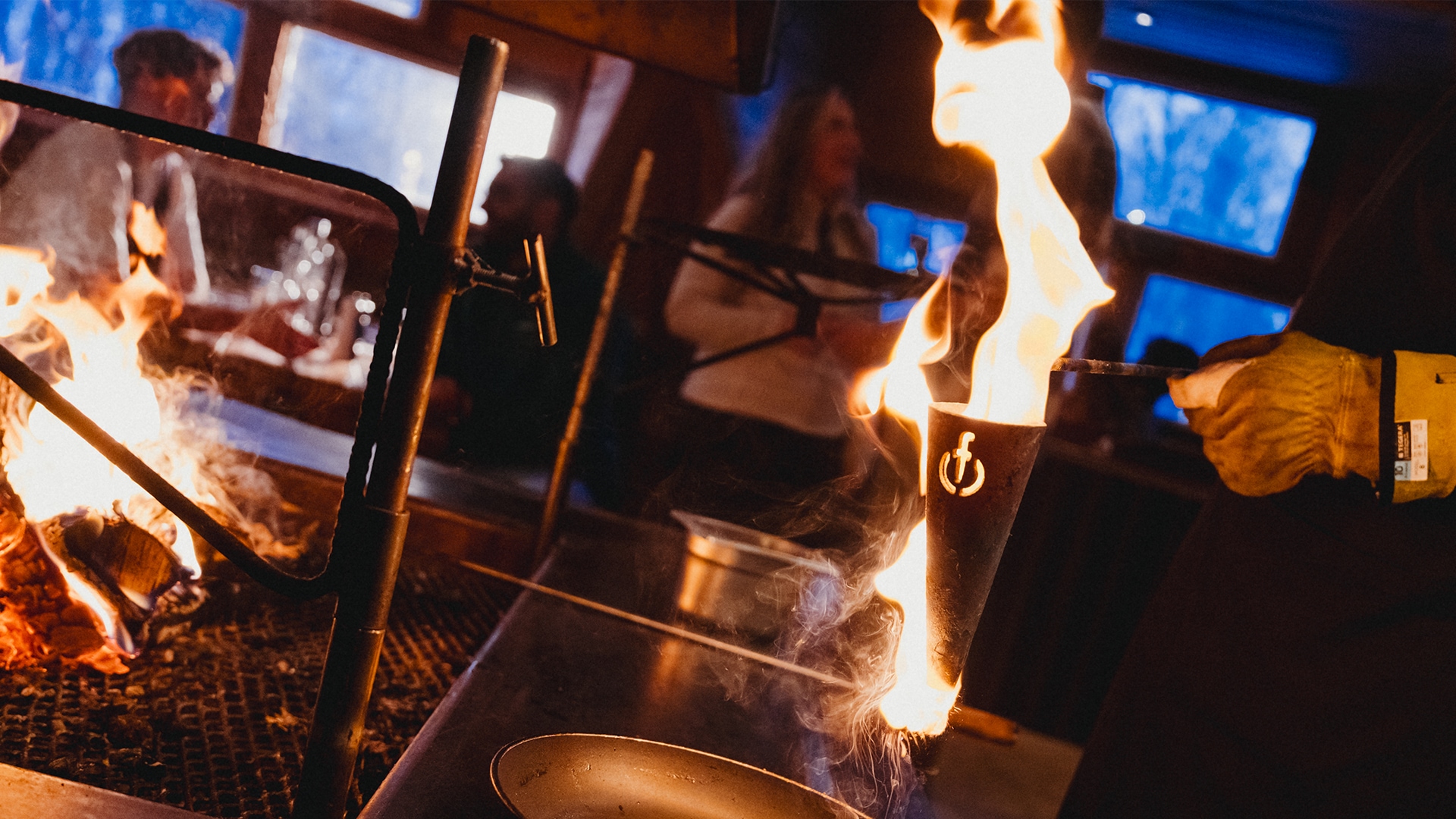 Flambiertes Essen über einer Feuerstelle in Innenräumen mit Menschen im Hintergrund