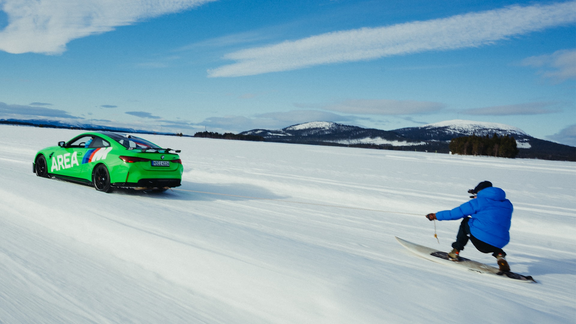BMW M grün mit AREA M Beschriftung in Seitenansicht zieht eine Person auf einem Snowboard über eine zugefrorene Fläche vor Berkette