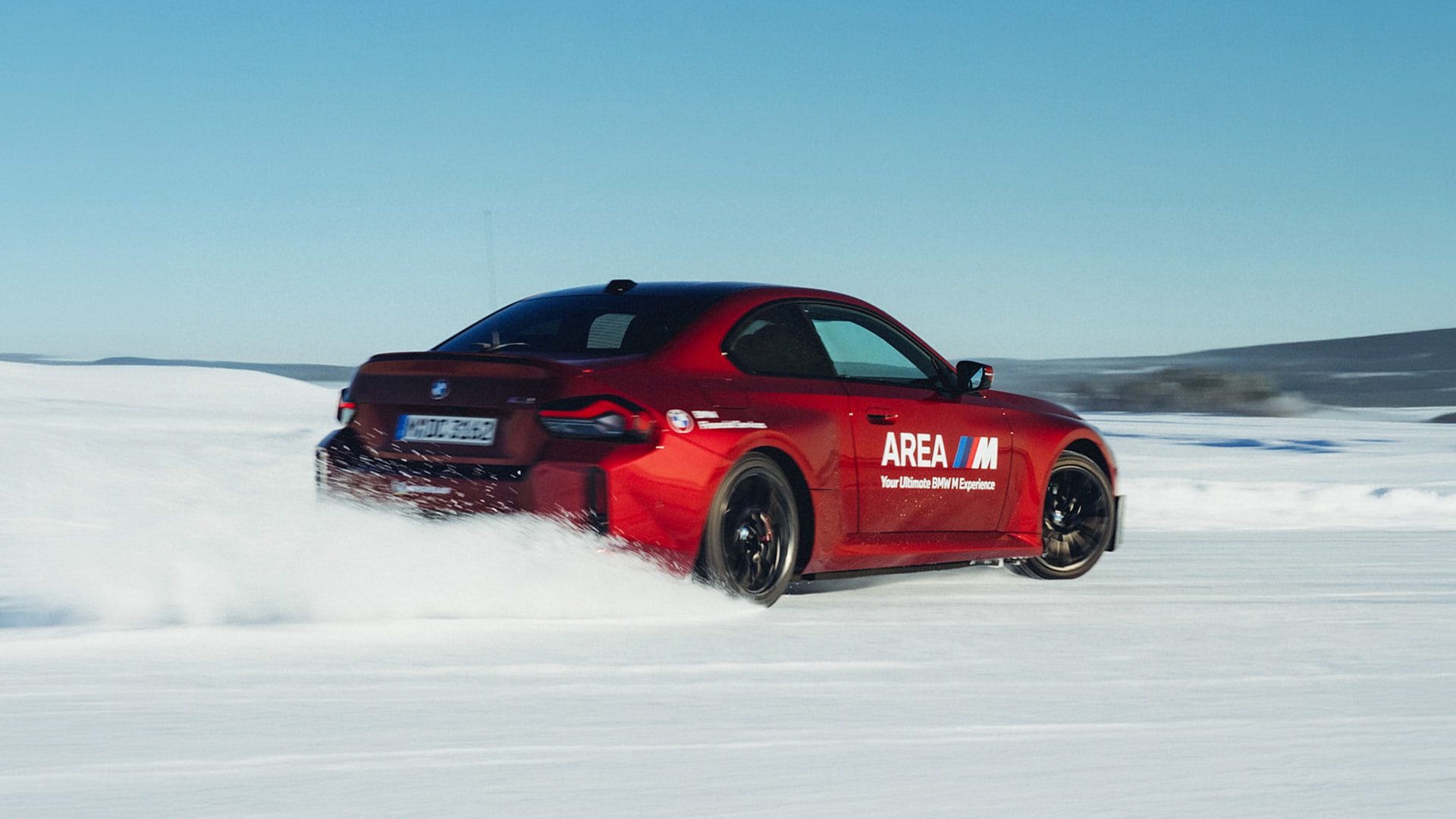 Driftender BMW M rot mit AREA M Beschriftung "Your Ultimate BMW M Experience" in dreiviertel Heckansicht im Schnee auf zugefrorener Fläche