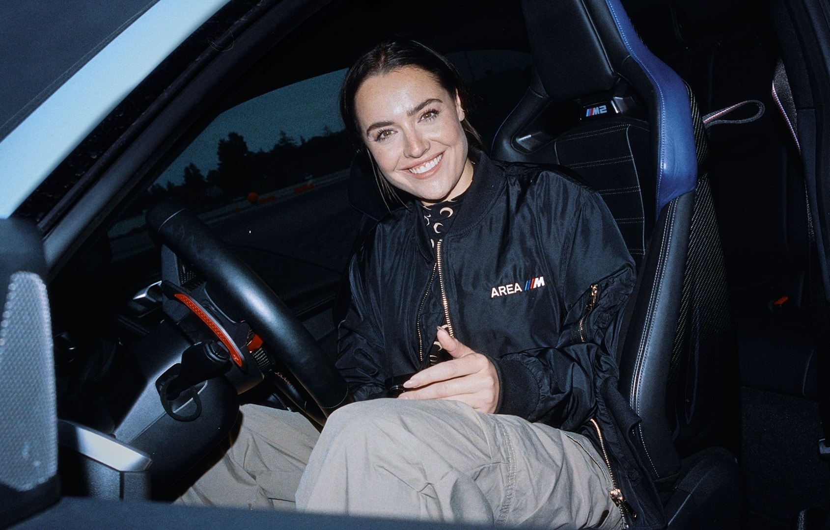 LächelndePerson mit AREA M Jacke sitzt im Rennsitz eines BMW M blau mit geöffneter Tür
