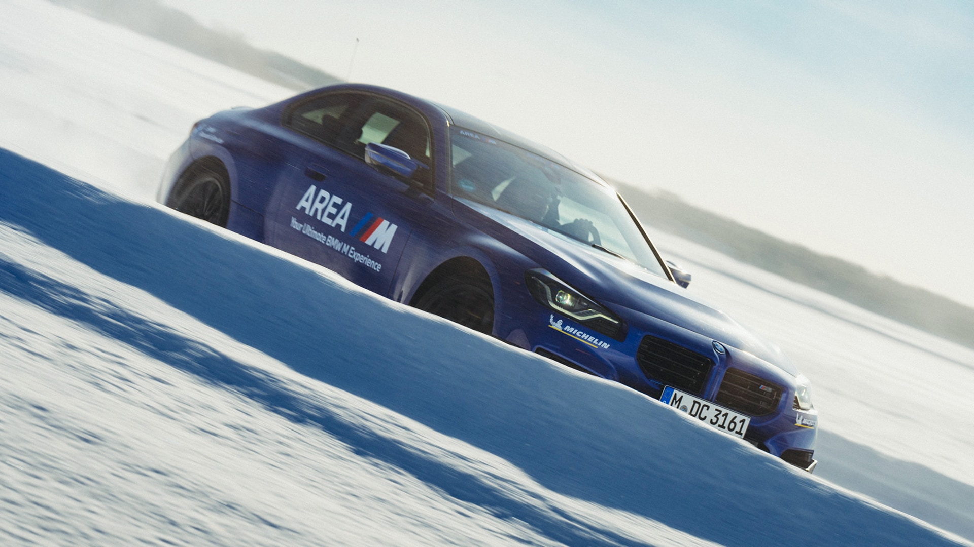 BMW M blau in dreiviertel Seitenansicht mit Area M Beschriftung „Your Ultimate BMW M Experience" und Michelin Partnerlogo auf Schnee