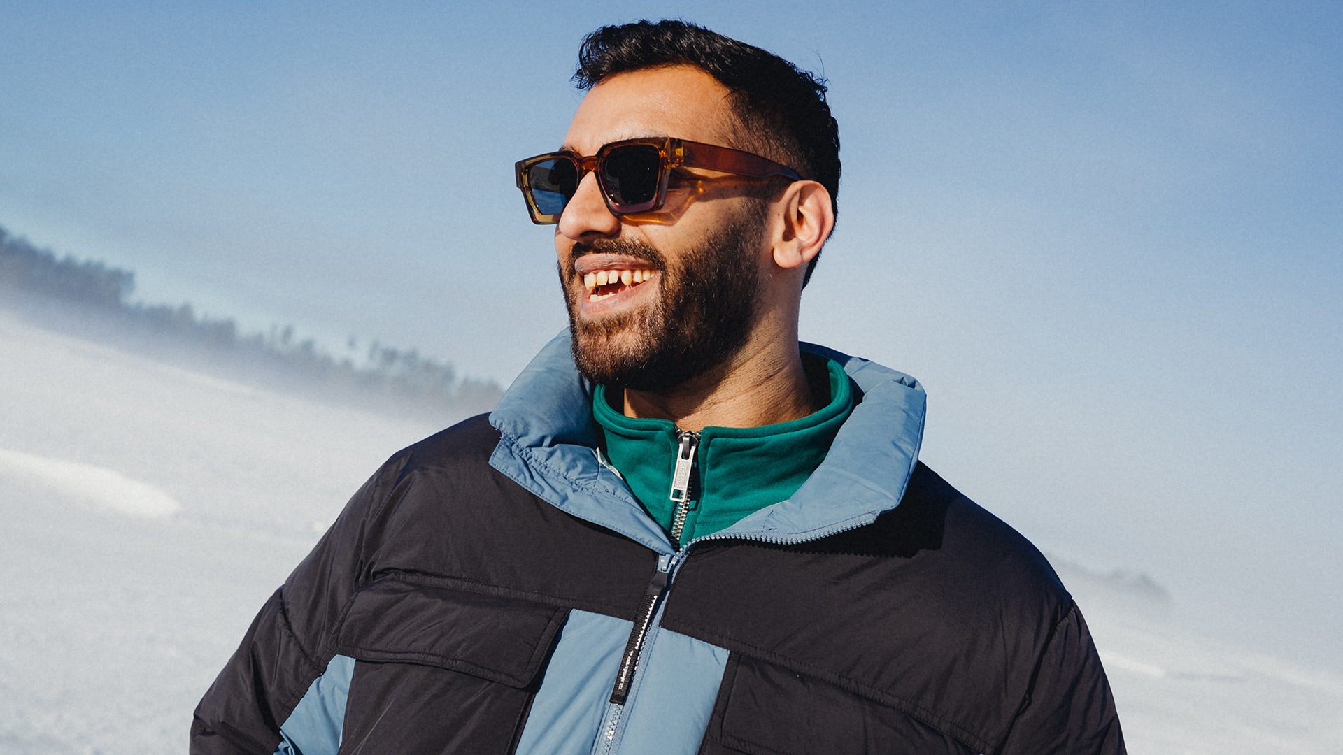 Lächelnde Person mit Sonnenbrille und Winterjacke blickt seitlich vor verschneiter Berglandschaft