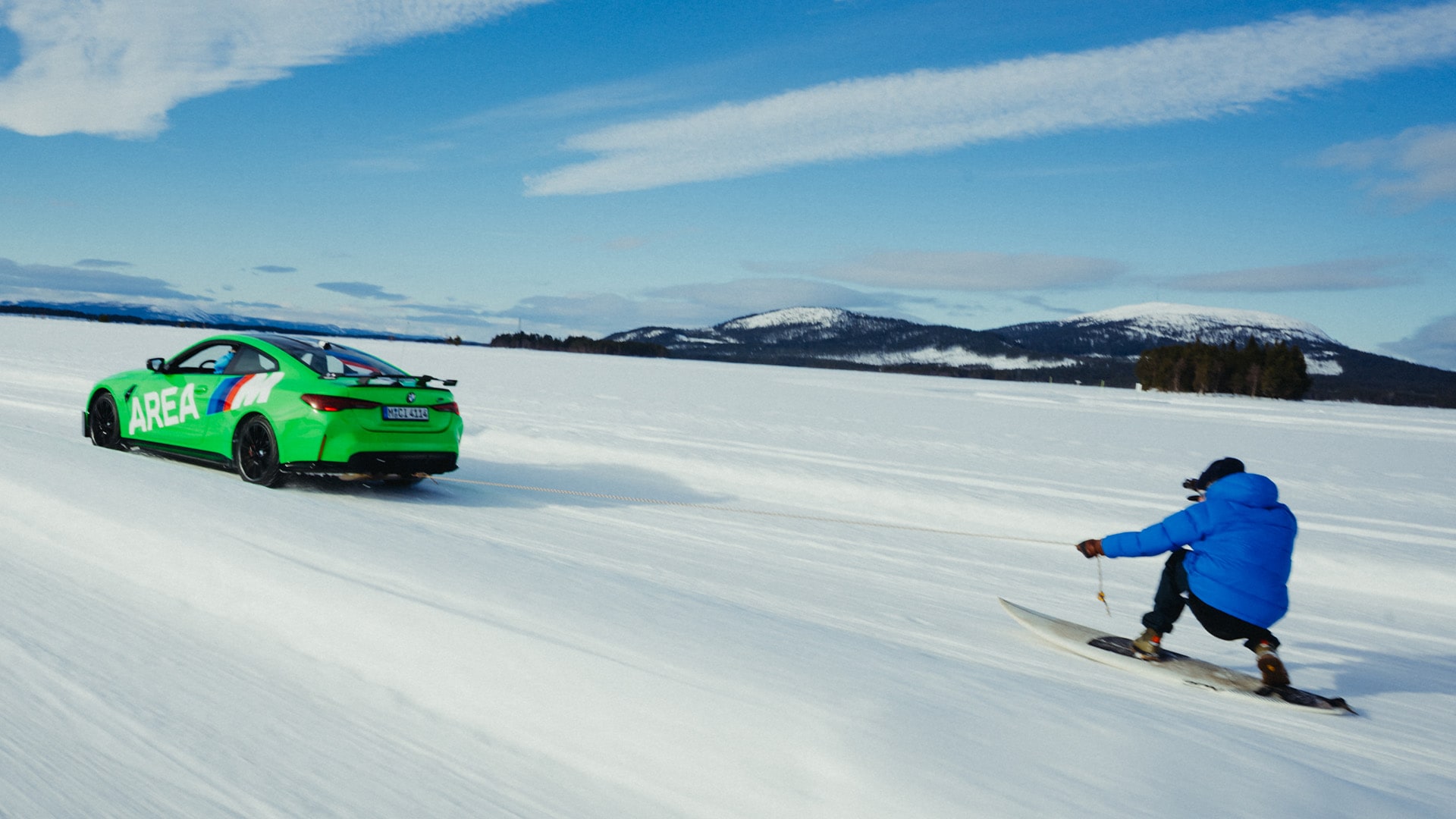 BMW M grün mit AREA M Beschriftung in Seitenansicht zieht eine Person auf einem Snowboard über eine zugefrorene Fläche vor Berkette