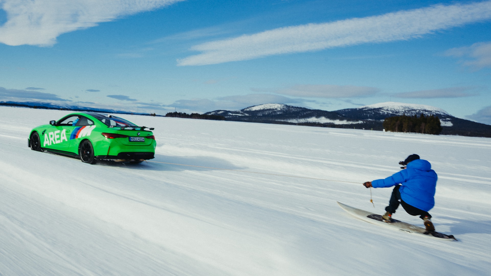BMW M grün mit AREA M Beschriftung in Seitenansicht zieht eine Person auf einem Snowboard über eine zugefrorene Fläche vor Berkette