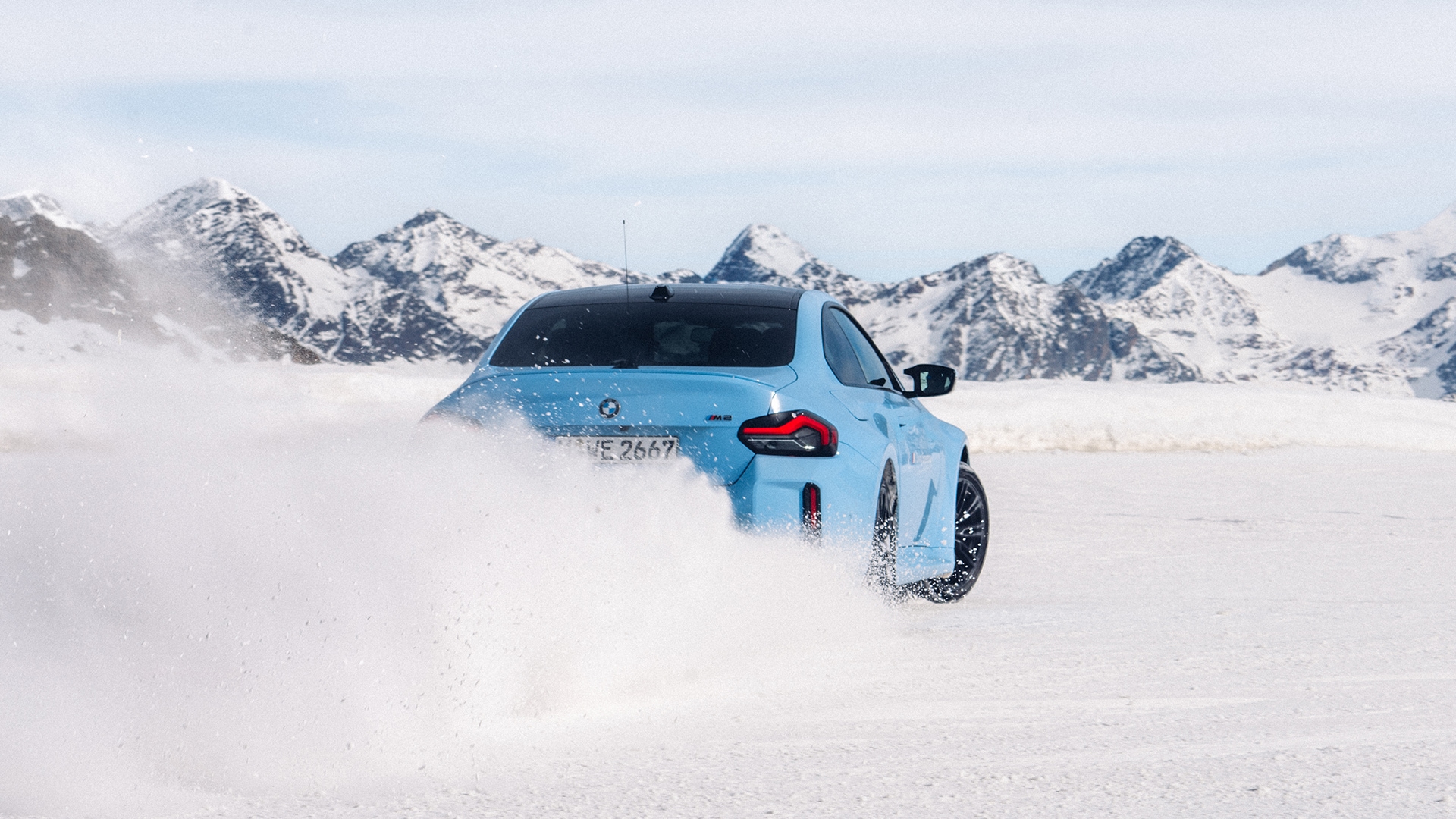 Driftender BMW M blau in Heckansicht im Schnee vor schneebedeckter Bergkette