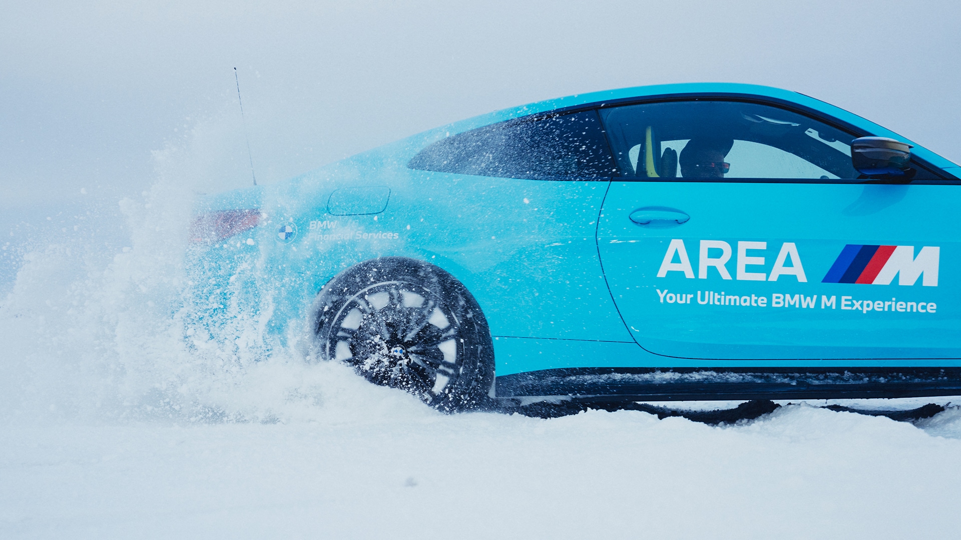 Türkisfarbenes BMW M mit AREA M Beschriftung „Your Ultimate BMW M Experience", seitliche Nahansicht mit starker Schneeaufwirbelung am Rad