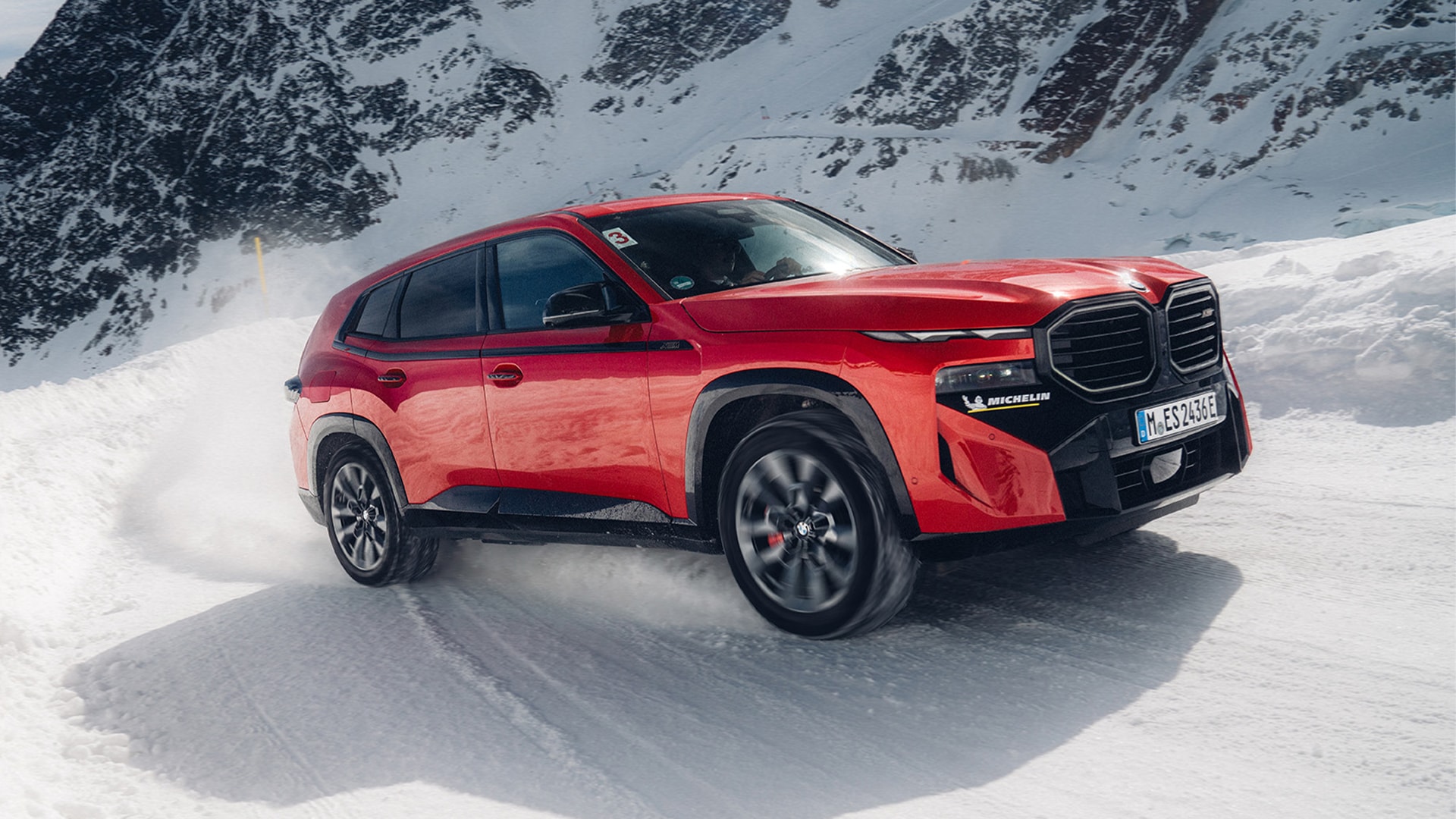 Rotes BMW XM im Drift, Dreiviertel-Frontansicht, Schneefontäne auf verschneiter Bergstraße vor Felsmassiv