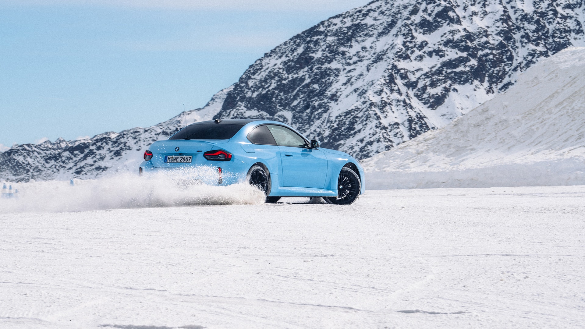 Driftender BMW M blau in Heckansicht im Schnee vor schneebedeckter Bergkette