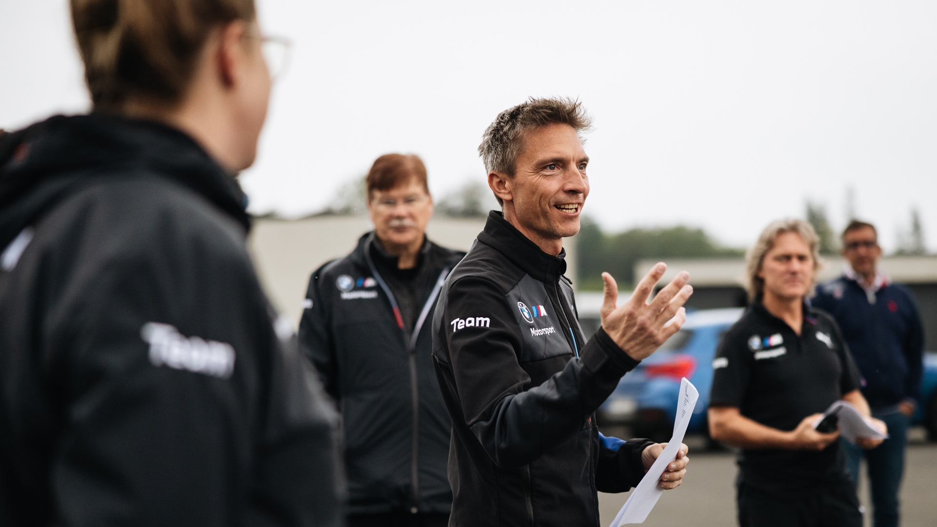 Eine Gruppe von Menschen in schwarzen Teamjacken mit BMW Motorsport-Branding, die sich im Freien versammelt haben. Eine Person in der Mitte gestikuliert während des Sprechens und hält Papiere in der Hand, anscheinend um Anweisungen oder ein Briefing zu geben. Mehrere Teammitglieder hören zu, in einer Umgebung im Freien mit Fahrzeugen im Hintergrund unter einem bewölkten Himmel.