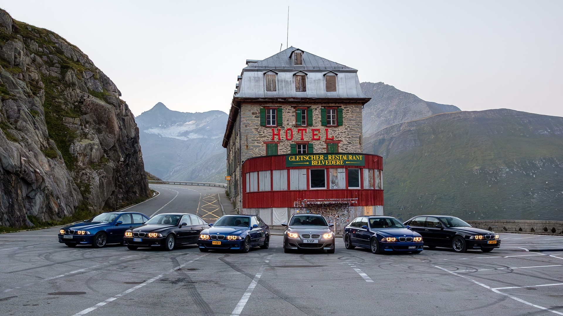 Eine Ansammlung von BMW M5 Fahrzeugen in einer Kehre vor einem Berghotel. 