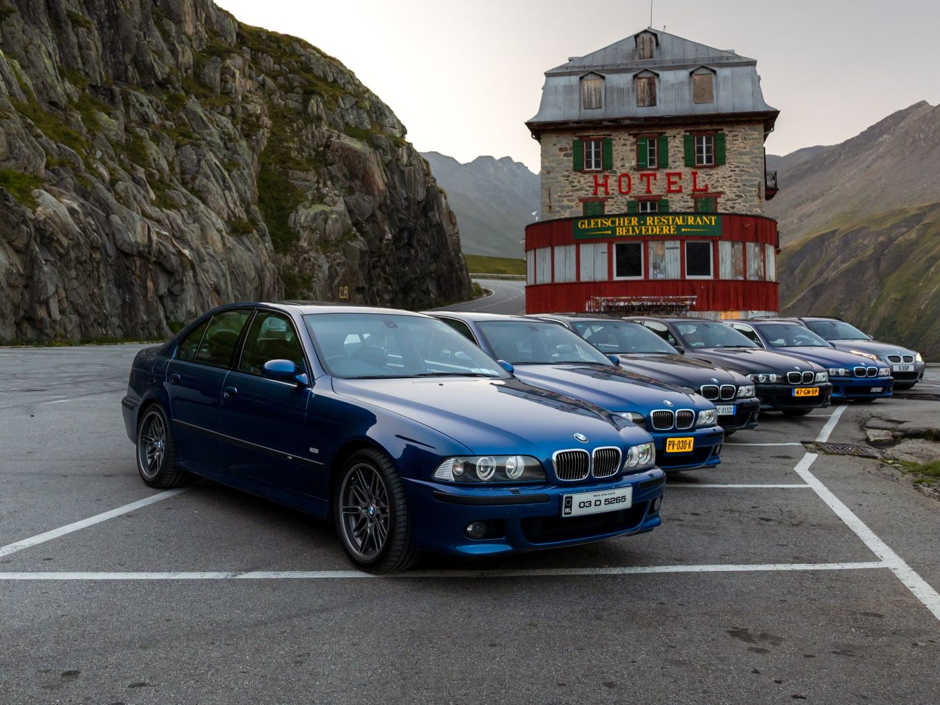 Eine Reihe älterer BMW M5 Modelle parkt in einer Kehre vor einem Berghotel. 