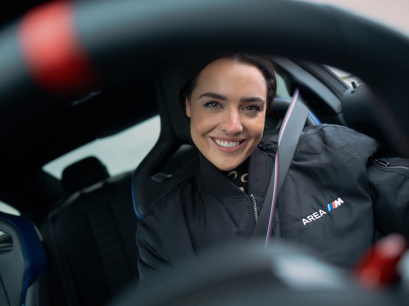 Becky Evans, strapped into a BMW M2, wearing an AREA M jacket. 